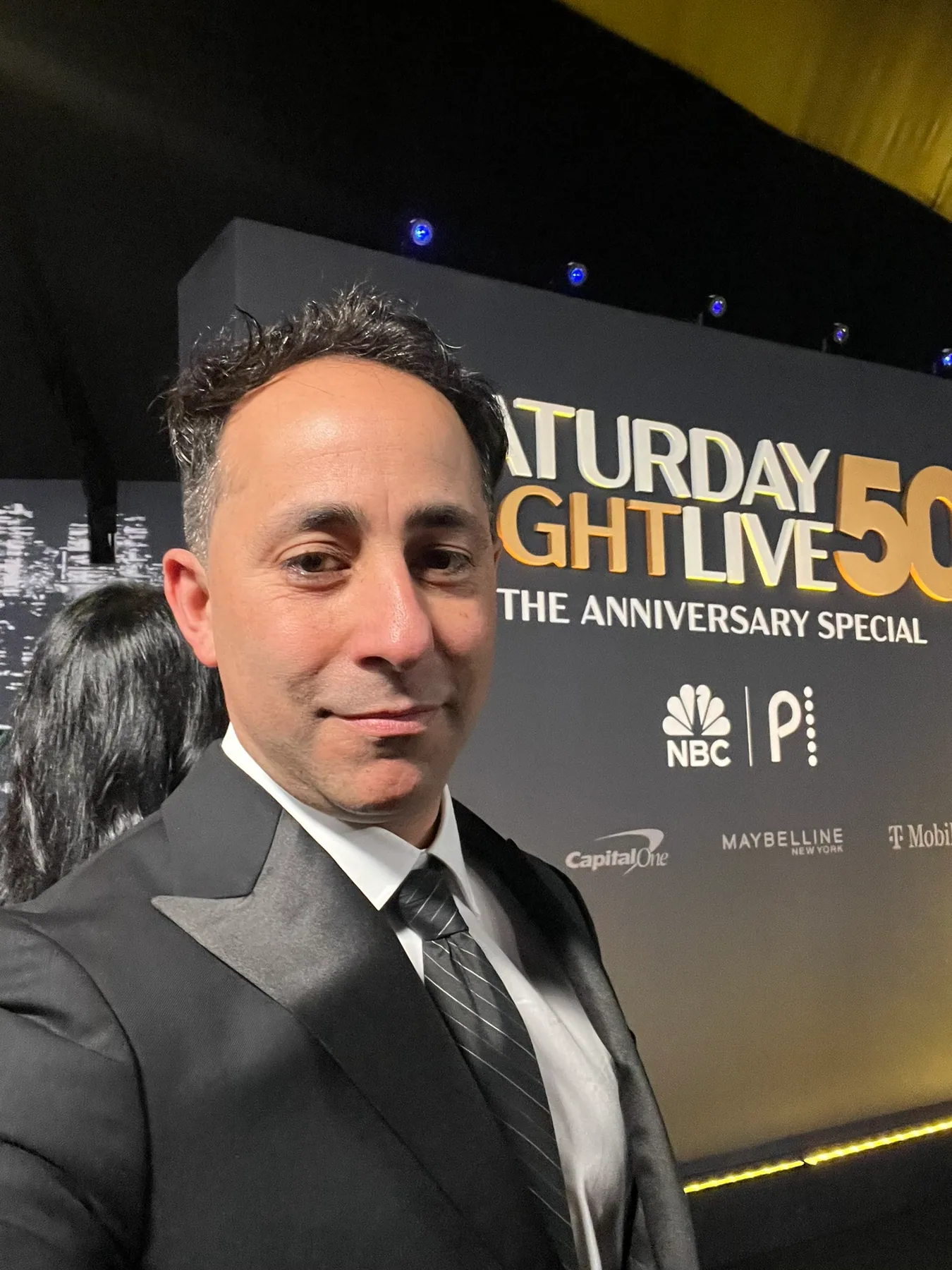 Matthew Legreca in a tuxedo with a handheld microphone at a red-carpet event backdrop